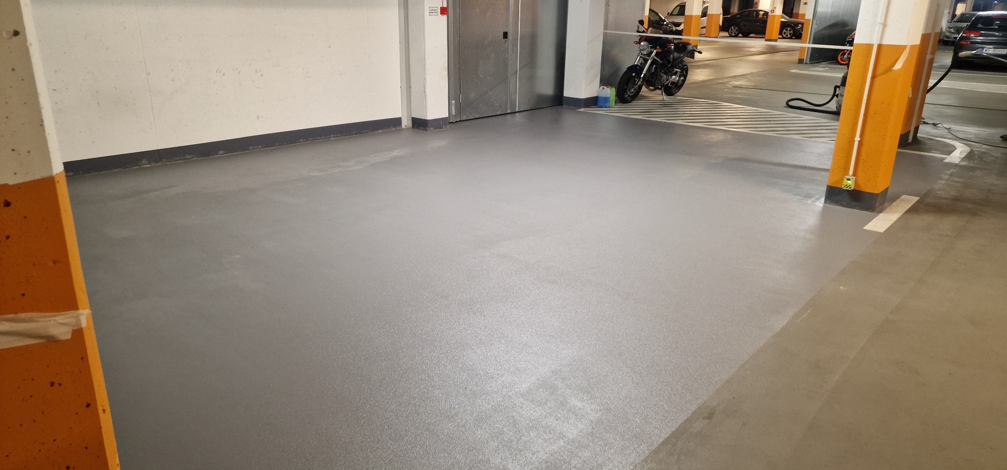 Szara, gładka posadzka w garażu podziemnym po renowacji, widoczny motocykl i zaparkowane samochody w tle, pomarańczowe słupy oddzielające miejsca parkingowe.