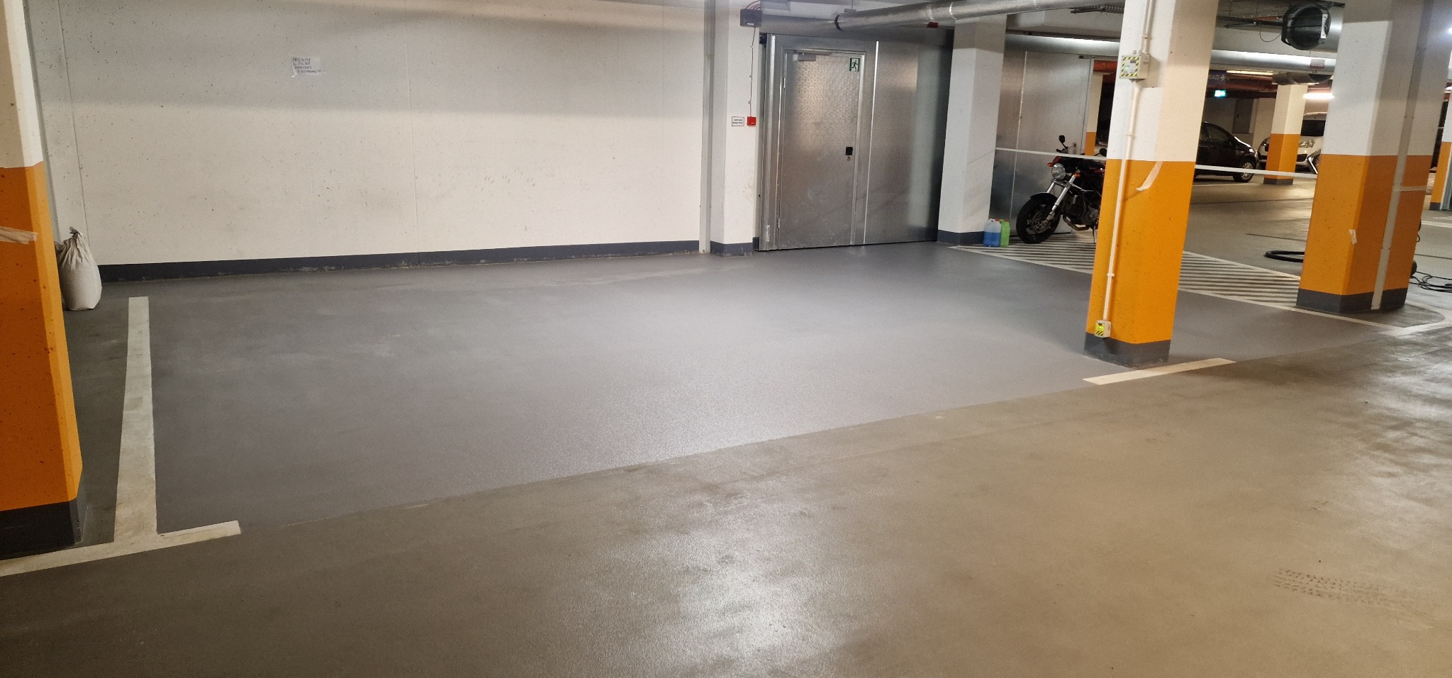 Szara posadzka żywiczna w garażu podziemnym, widoczne żółte słupy i motocykl w tle.