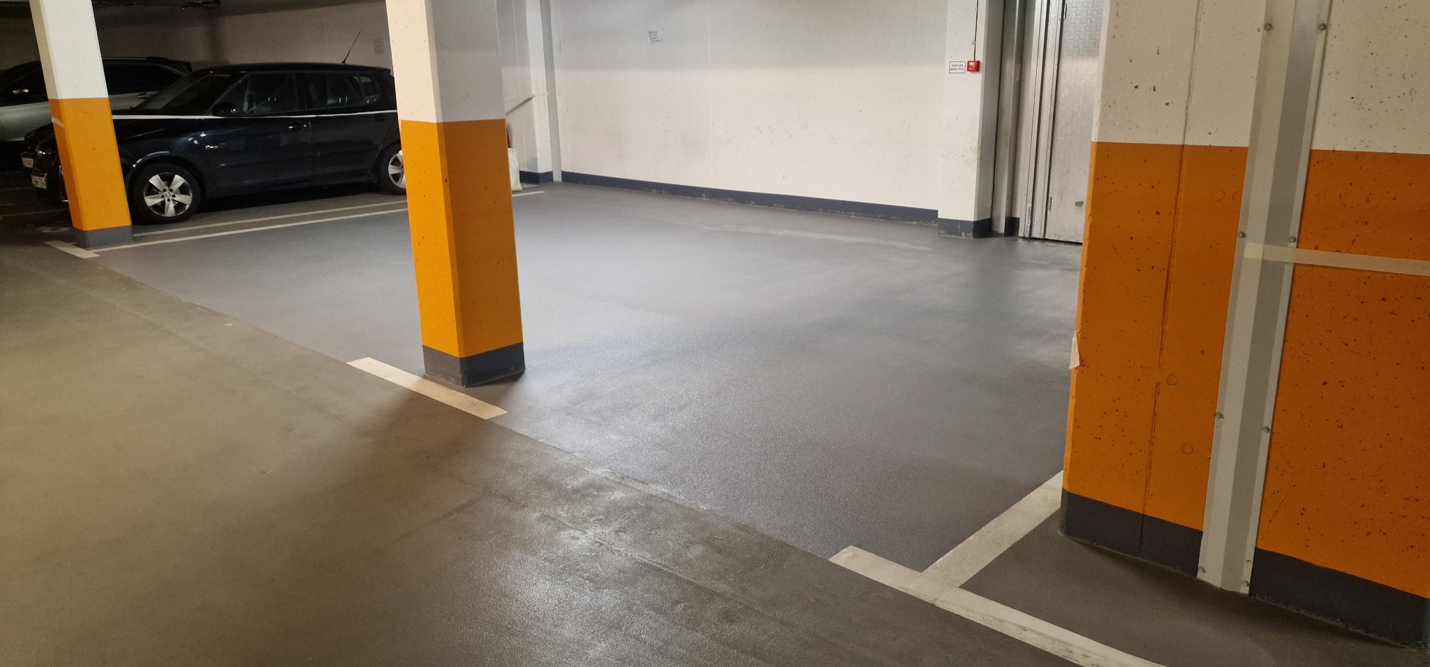 Szara, lśniąca posadzka w garażu podziemnym z pomalowanymi na żółto filarami i zaparkowanym samochodem w tle.