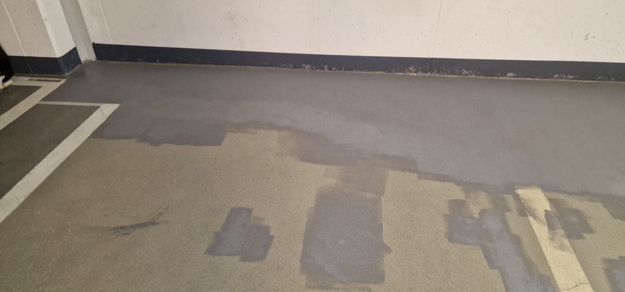 Fragment betonowej posadzki w trakcie wylewania żywicy, widoczne niedomalowane obszary i białe linie wyznaczające miejsca parkingowe.