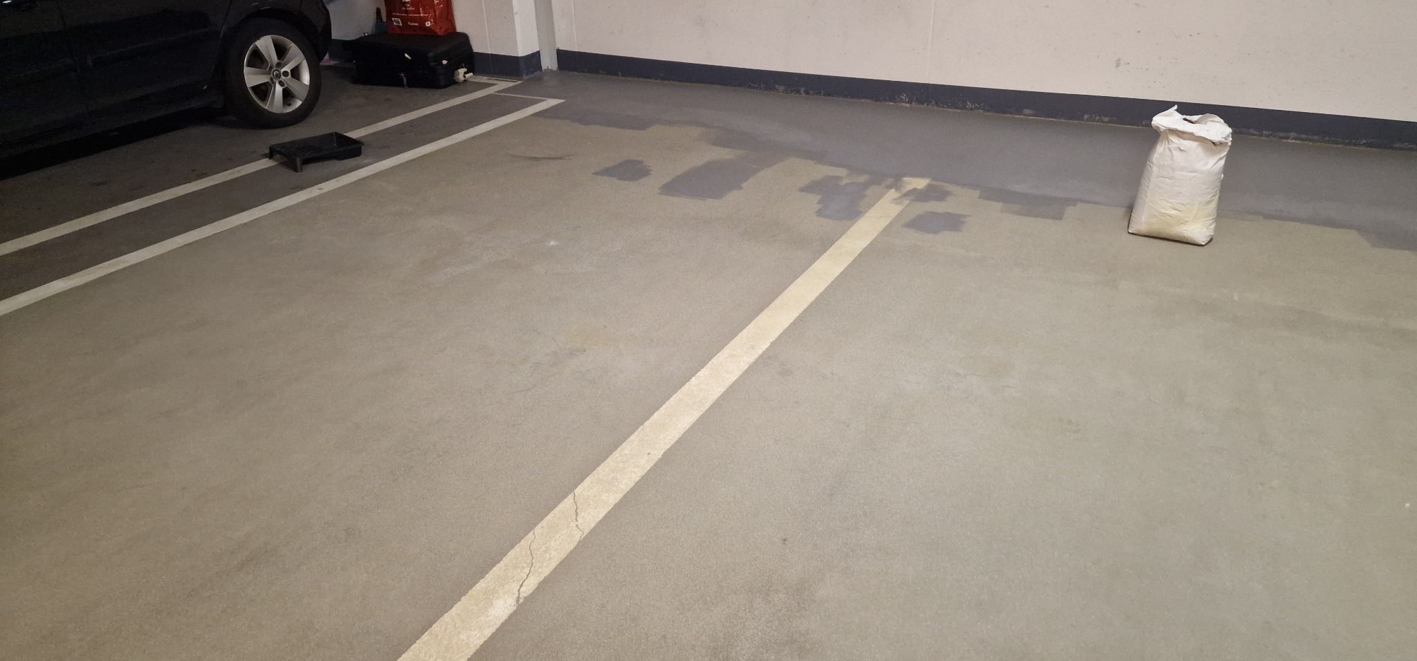Gładka, szara posadzka w garażu z wyznaczonymi białymi liniami stanowisk parkingowych, częściowo pokryta świeżo wylaną, ciemniejszą warstwą wyrównującą, obok zaparkowanego samochodu i worka...