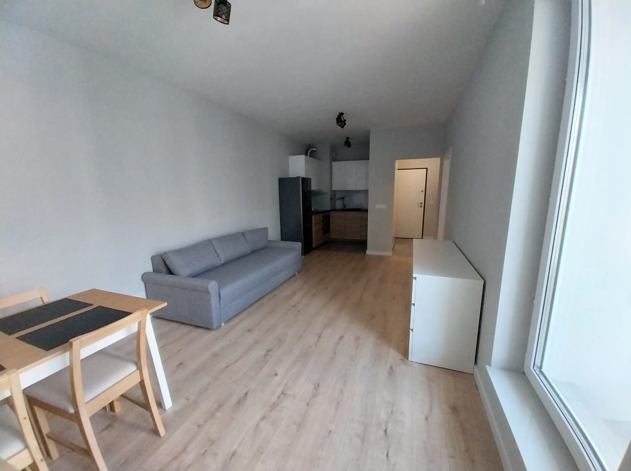 Wnętrze mieszkania po remoncie: jasne ściany, panele podłogowe, szara sofa, stół z krzesłami, biała komoda i widok na aneks kuchenny z czarną lodówką.