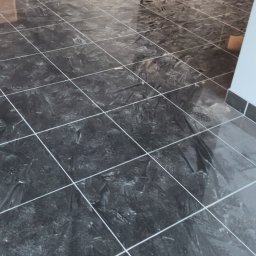 Clean Paradise - Błyszcząca podłoga z ciemnych, kwadratowych płytek imitujących marmur, z jasnymi fugami, widok na fragment ściany i drzwi w tle.