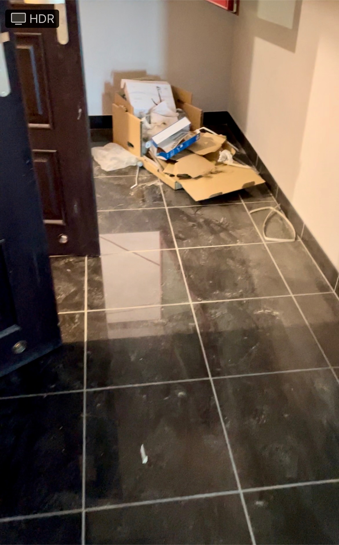 Korytarz z ciemnymi, błyszczącymi płytkami podłogowymi, na których leżą kartony po rozpakowanym sprzęcie i śmieci, widoczne przy otwartych drzwiach.