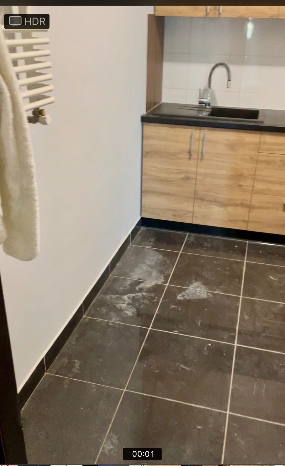 Fragment łazienki z czarną, częściowo zabrudzoną podłogą w płytkach, widoczna umywalka z szafką w tle oraz biały grzejnik drabinkowy z powieszonym ręcznikiem.