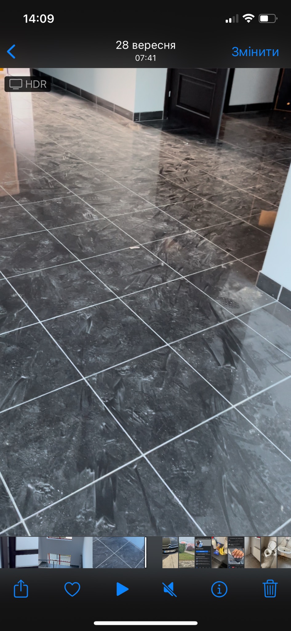 Błyszcząca podłoga z ciemnych, kwadratowych płytek imitujących marmur, z jasnymi fugami, widok na fragment ściany i drzwi w tle.