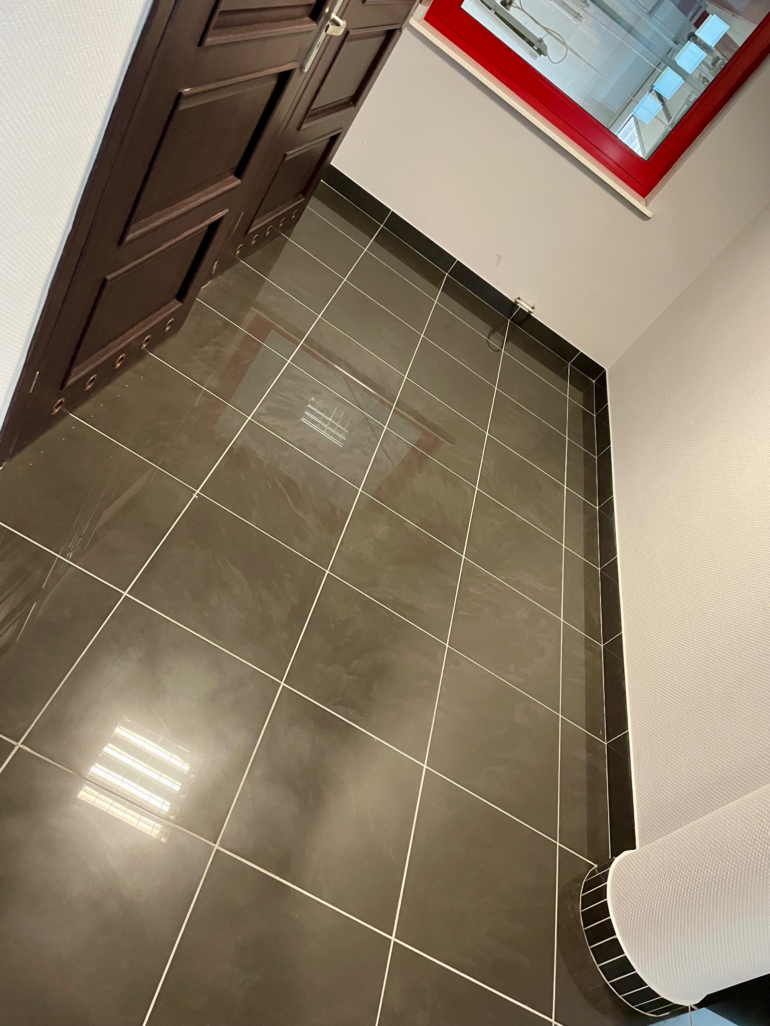 Błyszczące, ciemne płytki podłogowe z jasnymi fugami, widok z dołu w kierunku okna dachowego z czerwoną ramą i brązowych drzwi.