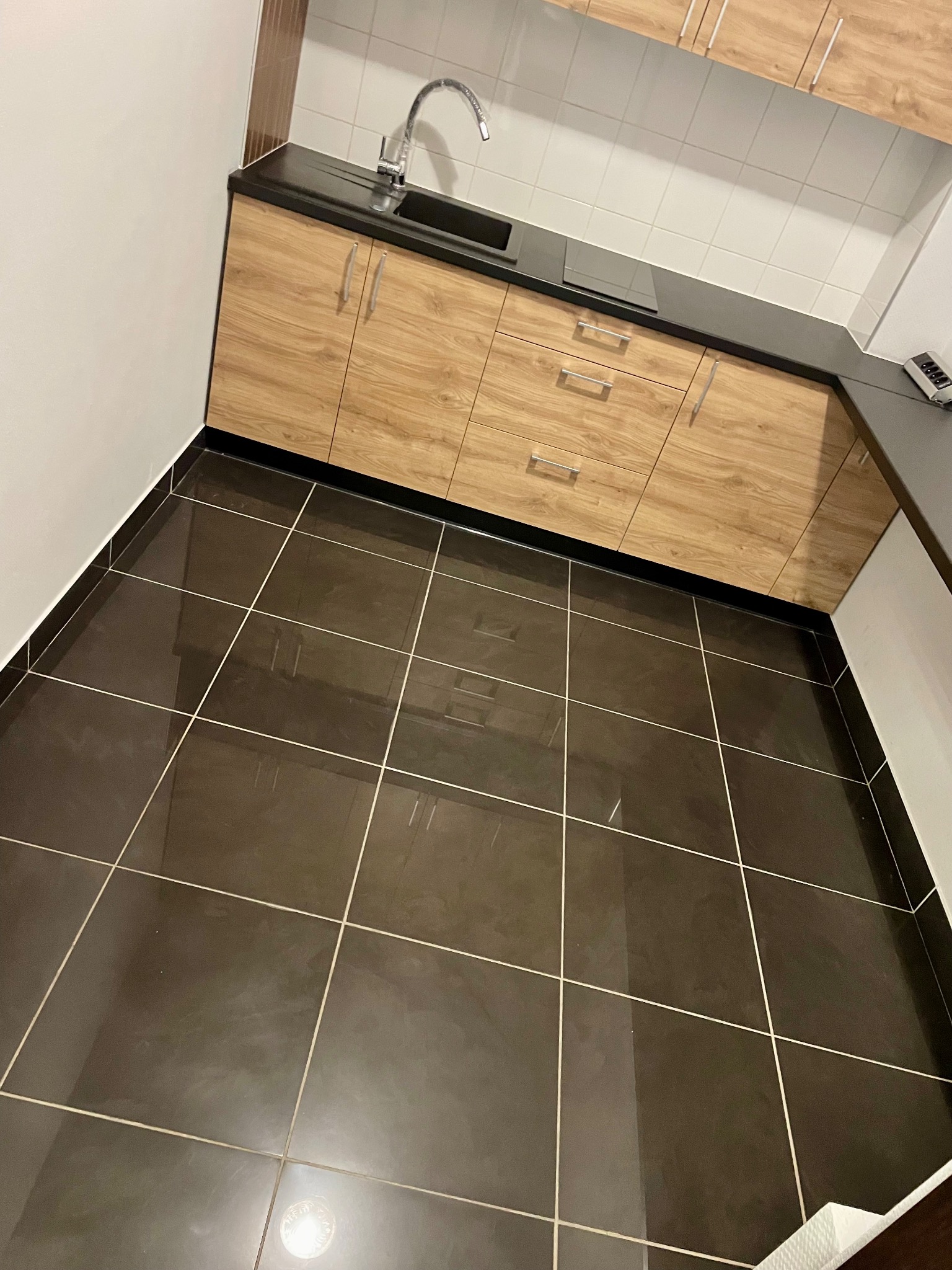 Ukośne ujęcie nowej kuchni z drewnopodobnymi szafkami, czarnym blatem i lśniącą, ciemną podłogą z płytek ceramicznych.