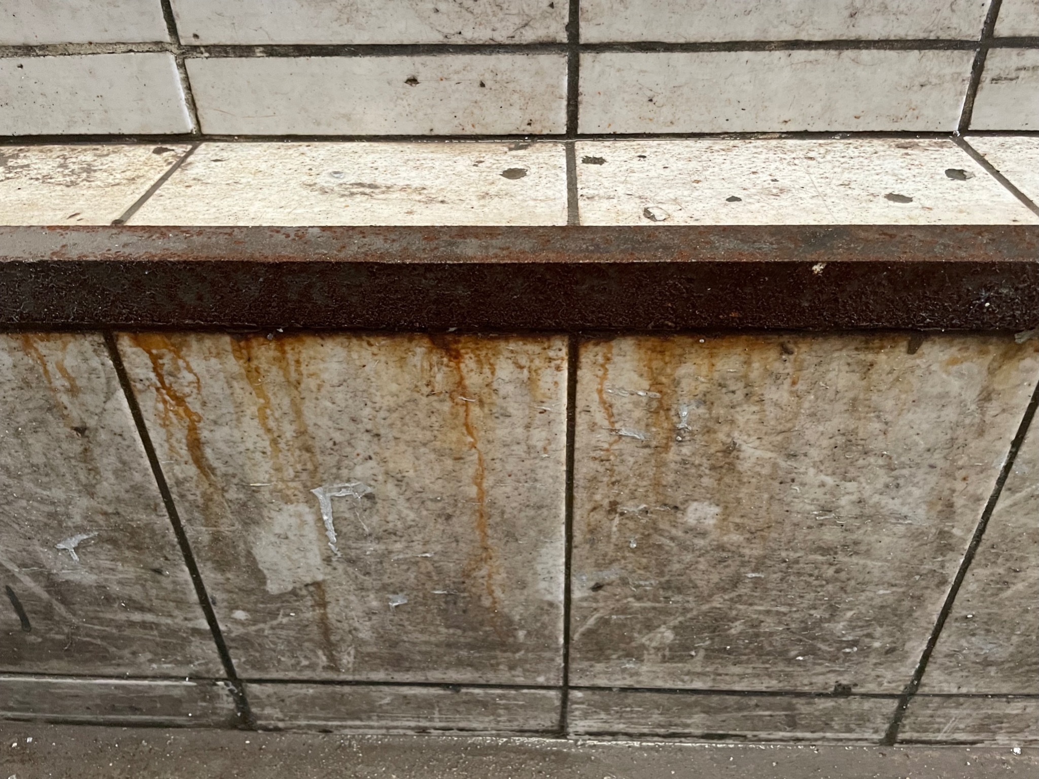 Zabrudzone, białe płytki ścienne z rdzawymi zaciekami i brudną, metalową listwą przypodłogową.