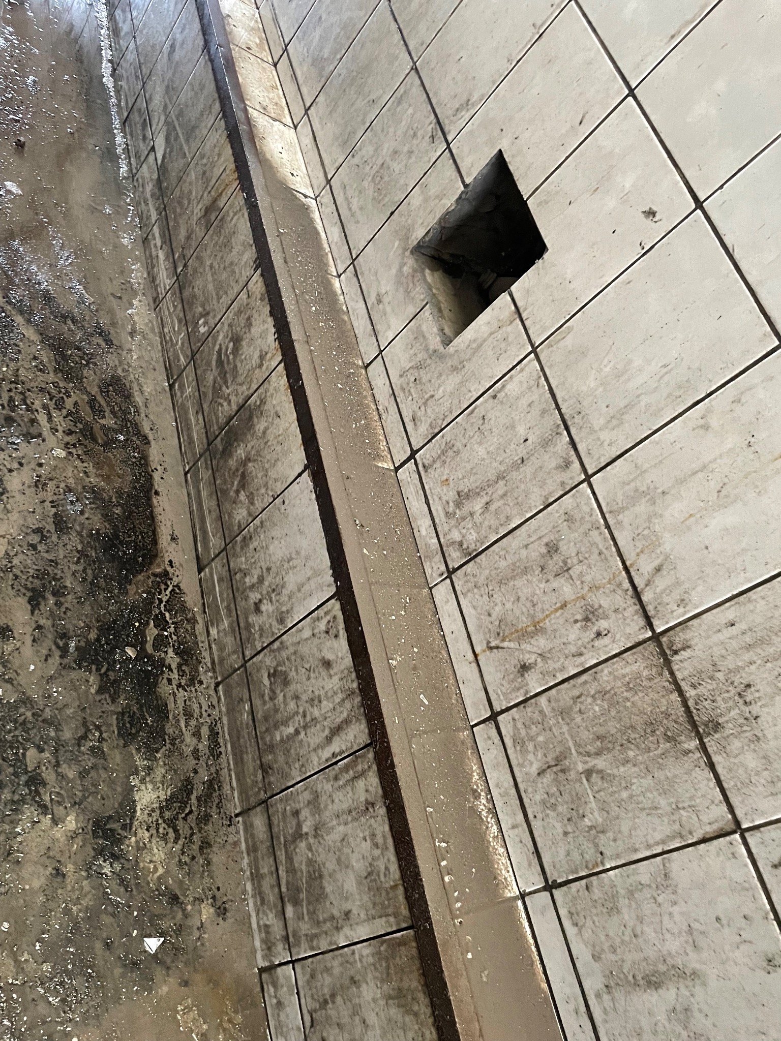 Zanieczyszczona podłoga wyłożona kwadratowymi płytkami ceramicznymi z widocznym odpływem kanalizacyjnym i resztkami brudnej wody.