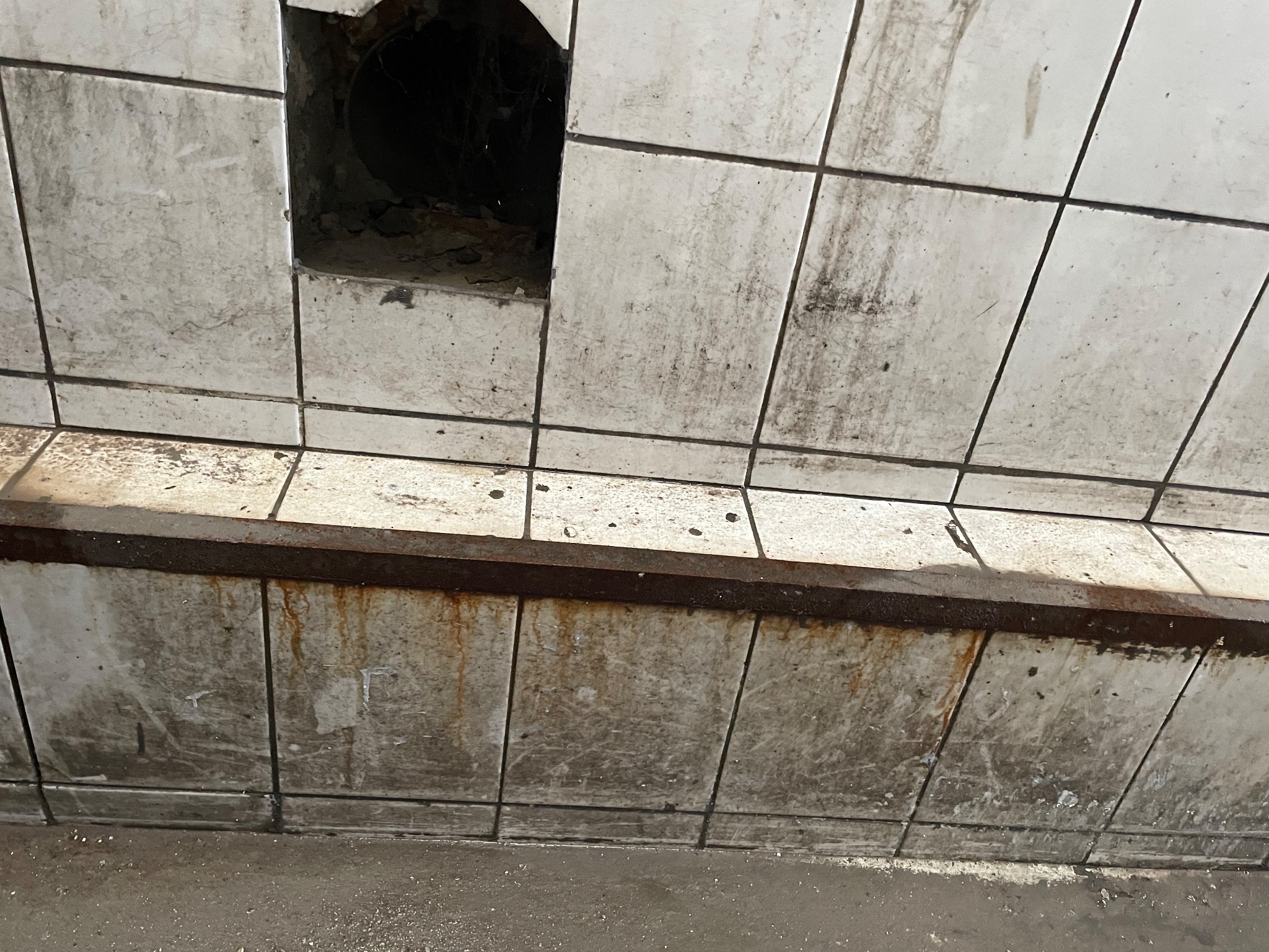 Zabrudzone, białe, kwadratowe płytki ścienne z ciemnymi fugami i śladami zacieków, z otworem w ścianie ukazującym wnętrze rury.