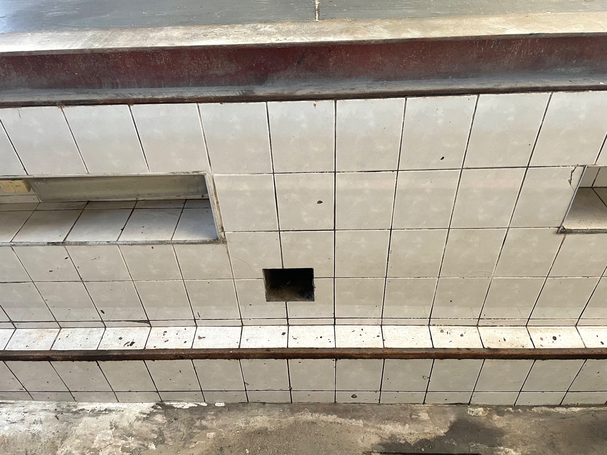 Brudne, białe kafelki ścienne z czarnymi plamami i kwadratową dziurą w środku, widoczne ubytki fugi i zabrudzenia.