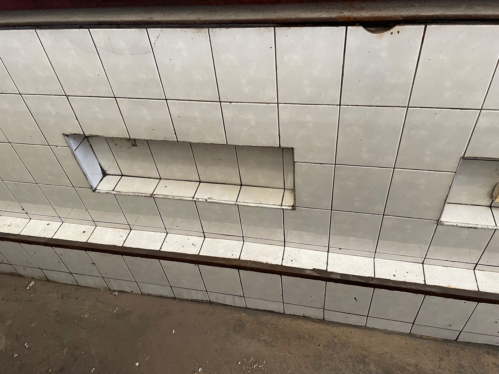 Białe, kwadratowe płytki ścienne z fugami, z wnęką w ścianie, widoczne zabrudzenia i osad.