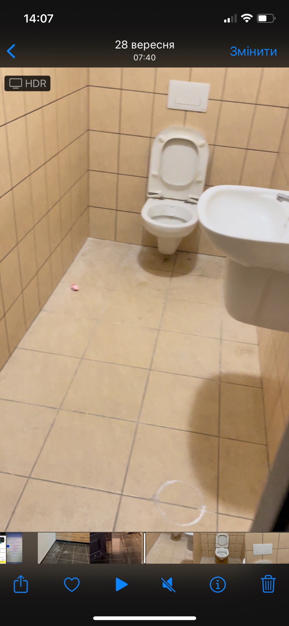 Toaleta z beżowymi płytkami, widoczne ślady po czyszczeniu na podłodze, otwarte siedzisko toalety, mała plamka na podłodze.