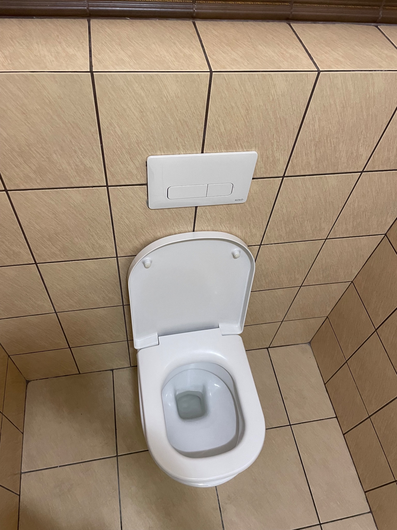 Biała toaleta marki Koło z otwartą klapą, widok z góry na tle beżowych płytek