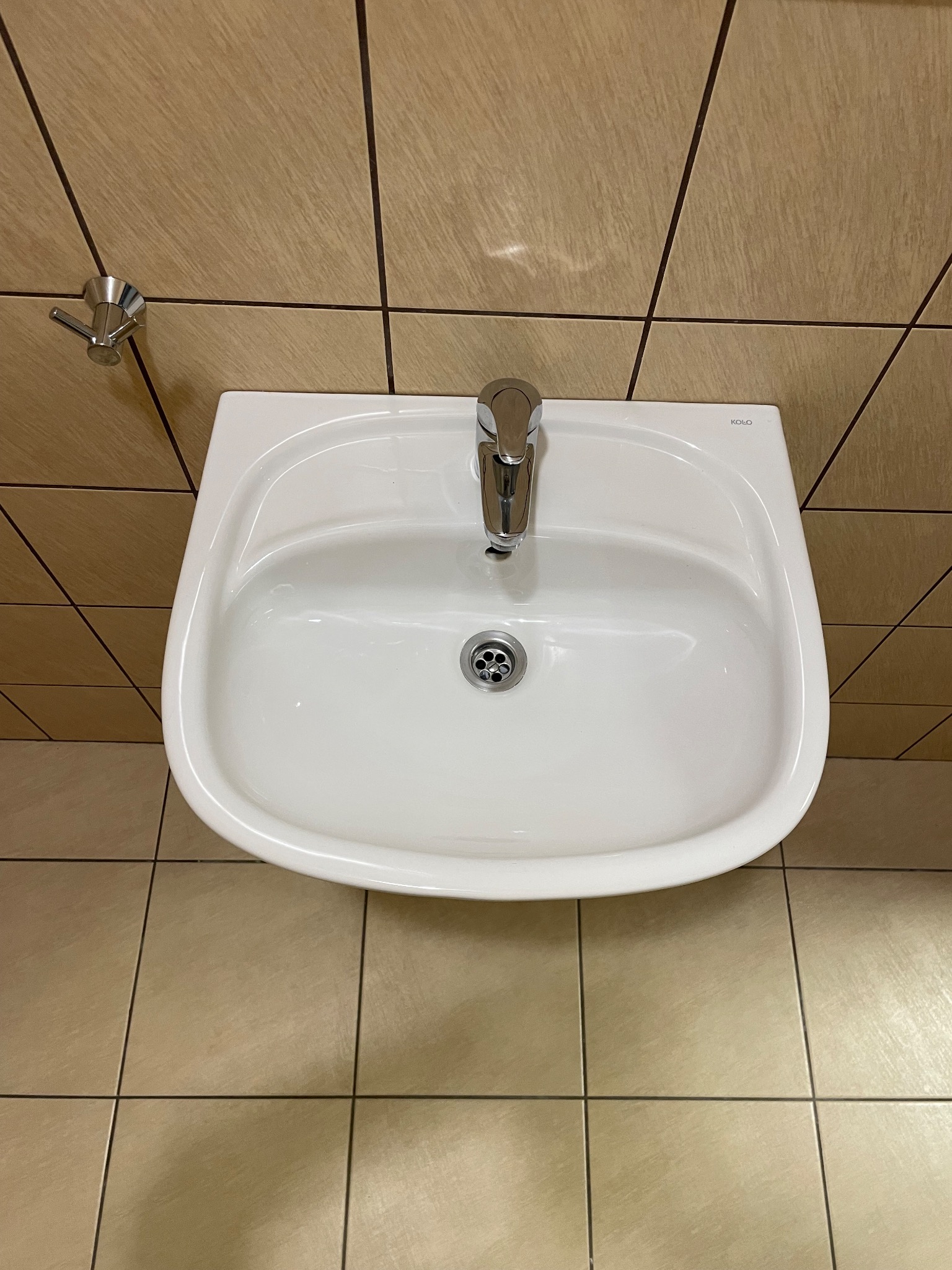 Biała umywalka marki Koło z chromowaną baterią i odpływem, zamontowana na tle beżowych płytek ściennych i podłogowych.