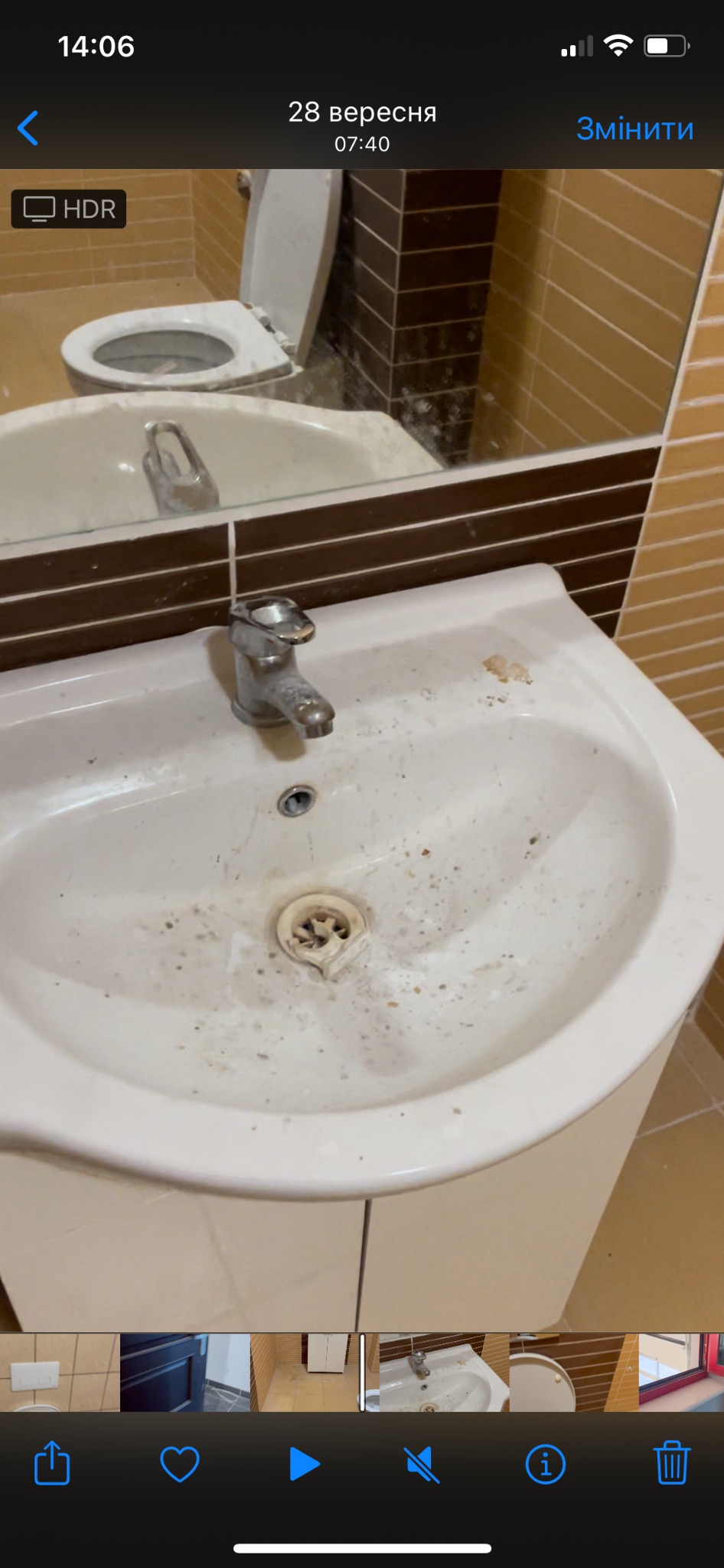 Zabrudzona biała umywalka z chromowaną baterią w łazience z brązowo-beżowymi płytkami, widoczne plamy i osady na powierzchni umywalki i w odpływie, odbicie sedesu w lustrze.