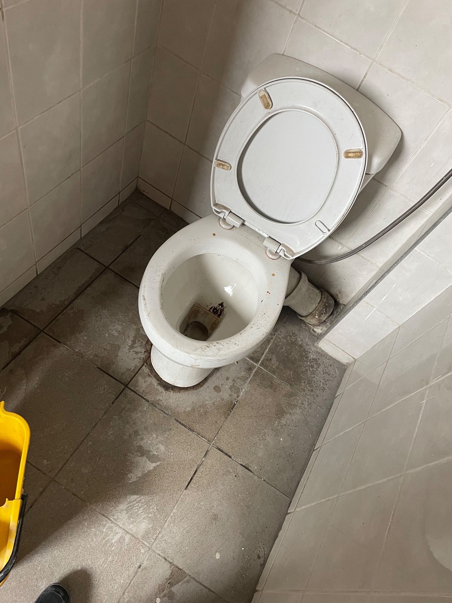 Zanieczyszczona toaleta z otwartą klapą, widoczne plamy na podłodze i ścianach z płytek, żółte wiadro w rogu kadru sugerujące potrzebę gruntownego czyszczenia.