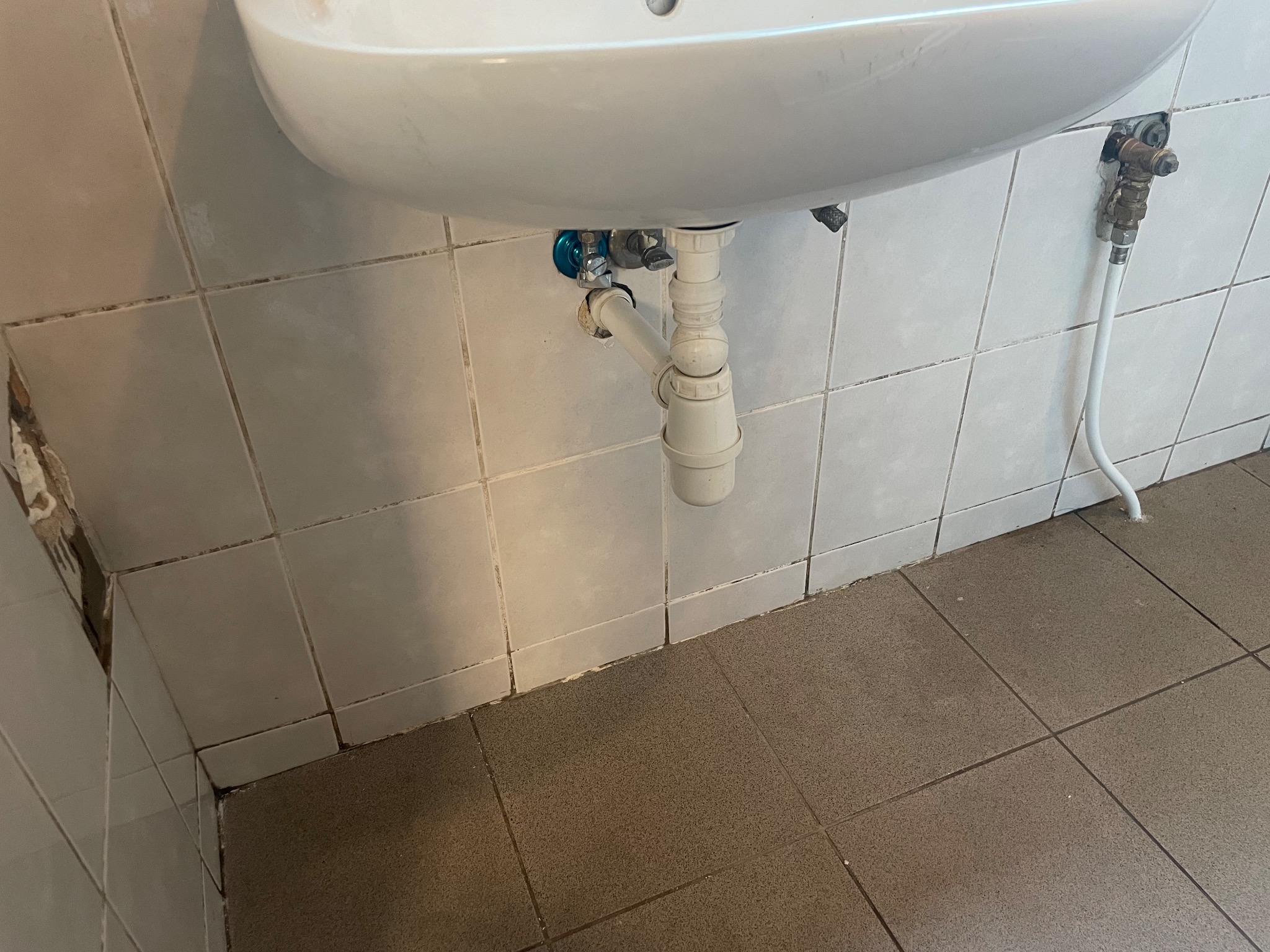 Biała umywalka z widoczną instalacją hydrauliczną, płytki ścienne z widocznymi zabrudzeniami fug, uszkodzony narożnik ściany, brudna podłoga.