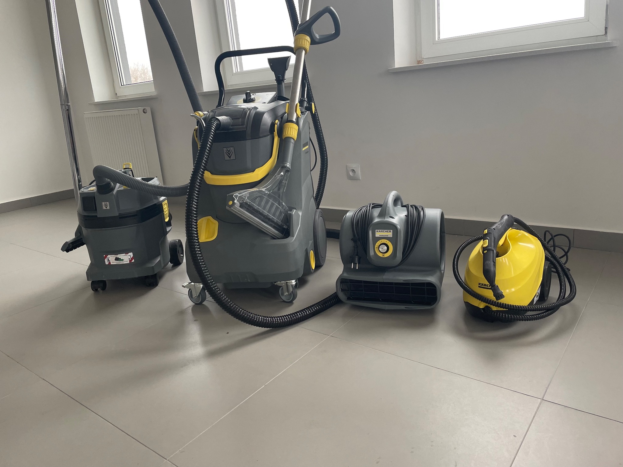 Profesjonalny sprzęt do czyszczenia: odkurzacz piorący z przezroczystą dyszą, odkurzacz przemysłowy i wentylator osuszający marki Kärcher, ustawione na szarej podłodze w jasnym pomieszczeniu z oknami.