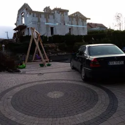 Niedokończony budynek z białego betonu komórkowego z drewnianą konstrukcją dachu, widoczna drabina, plac zabaw dla dzieci i czarny samochód osobowy na posesji z kostki brukowej.