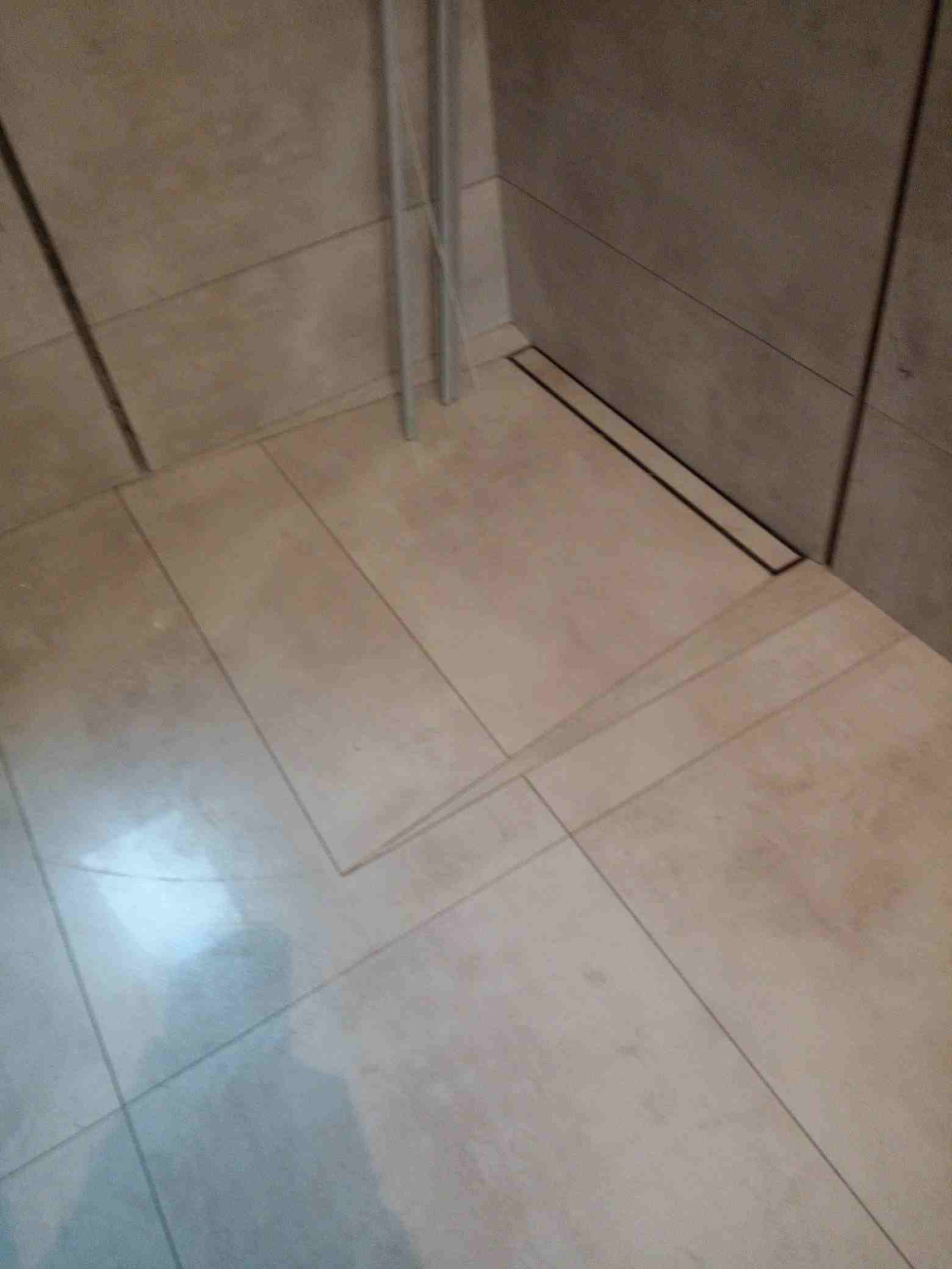 Ułożone płytki podłogowe w łazience z odpływem liniowym przy ścianie, widoczne fugi i fragment rur.