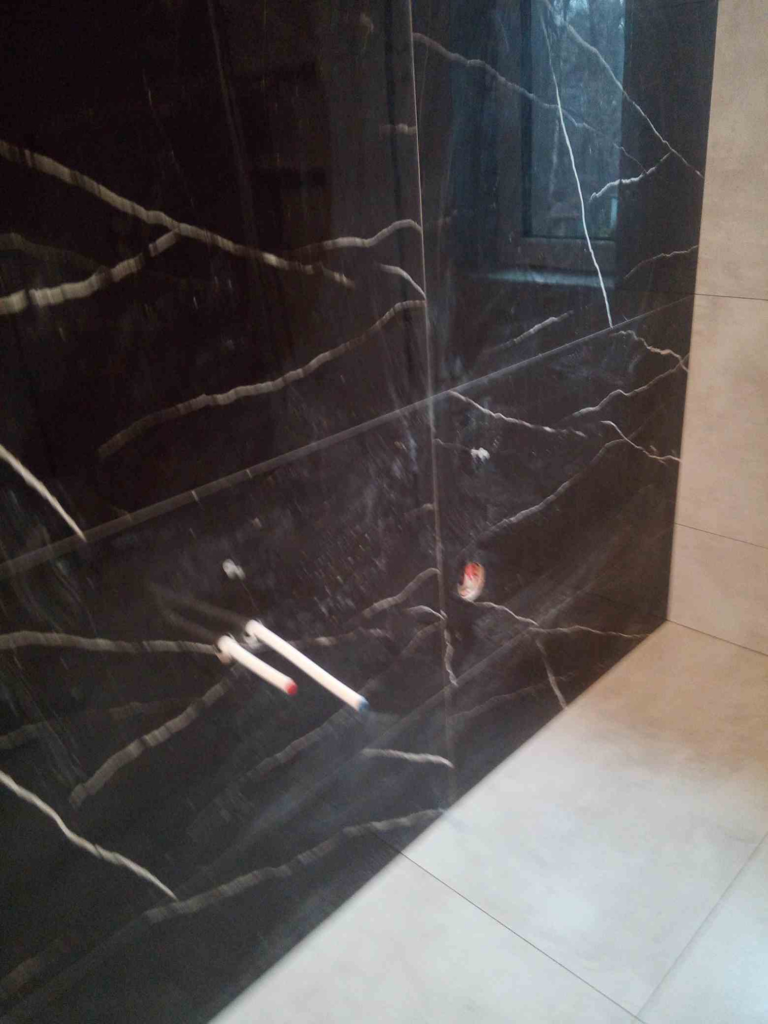 Ciemna, marmurkowa ściana z widocznymi rurami instalacji wodnej, kontrastująca z jasnymi płytkami podłogowymi, ukazująca etap prac wykończeniowych w łazience.