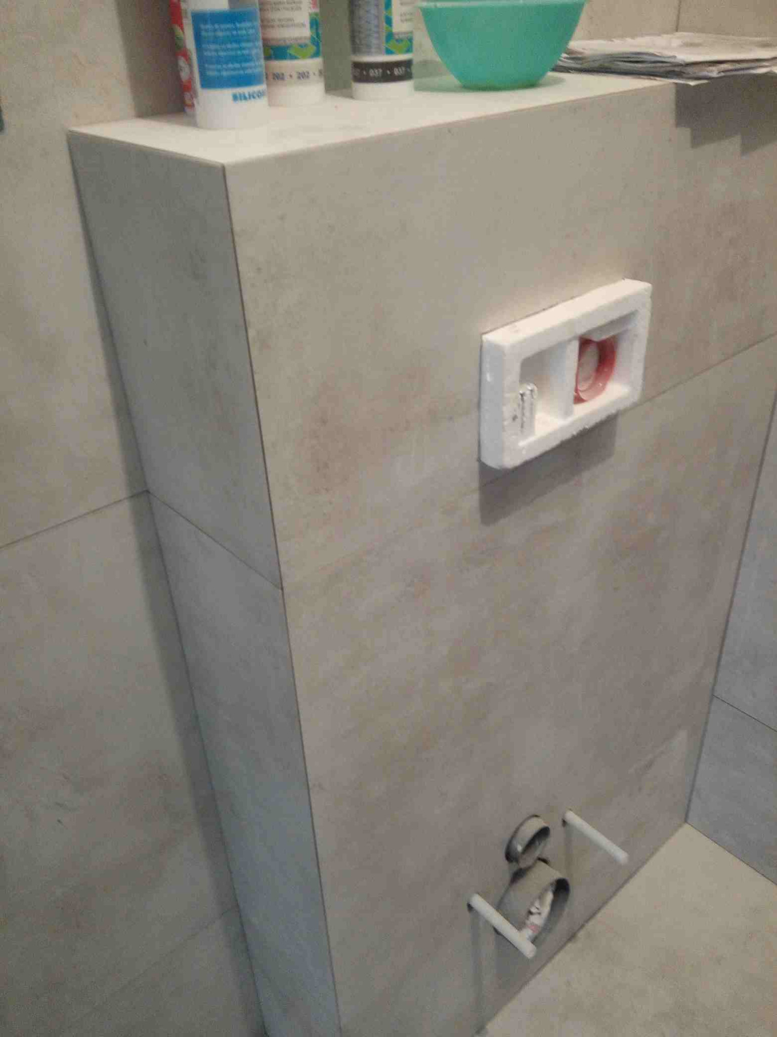 Obudowa stelaża podtynkowego WC wykończona płytkami, z widocznymi przyłączami kanalizacyjnymi i elementami montażowymi spłuczki.