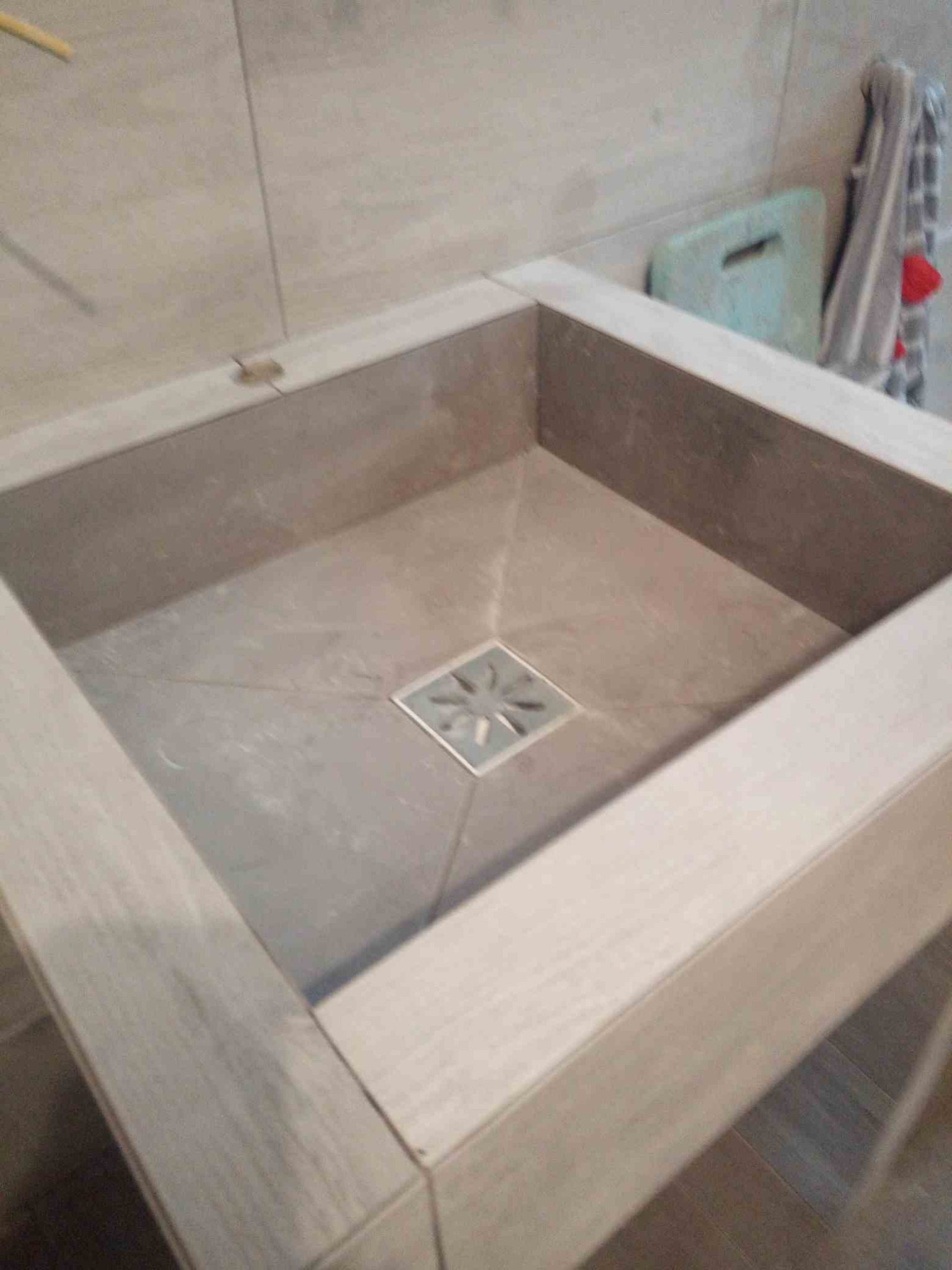 Wykończony brodzik prysznicowy z jasnoszarych płytek z odpływem liniowym w kształcie kwadratu z motywem kwiatowym, otoczony ramą z płytek imitujących drewno.