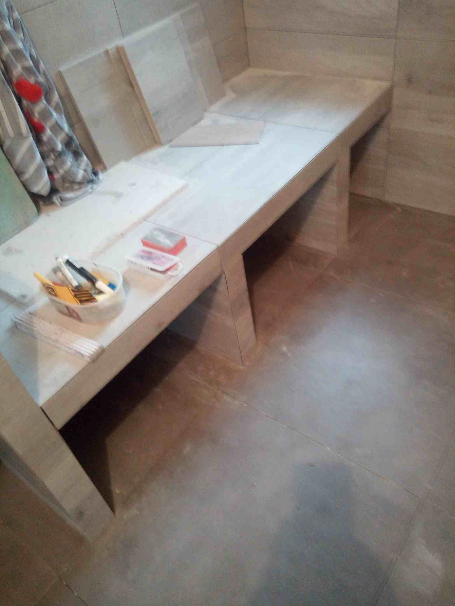 Wnętrze łazienki w trakcie prac glazurniczych: obudowany, wykafelkowany fragment ściany z półką, widoczne narzędzia i akcesoria w pojemniku, częściowo ułożone płytki podłogowe.