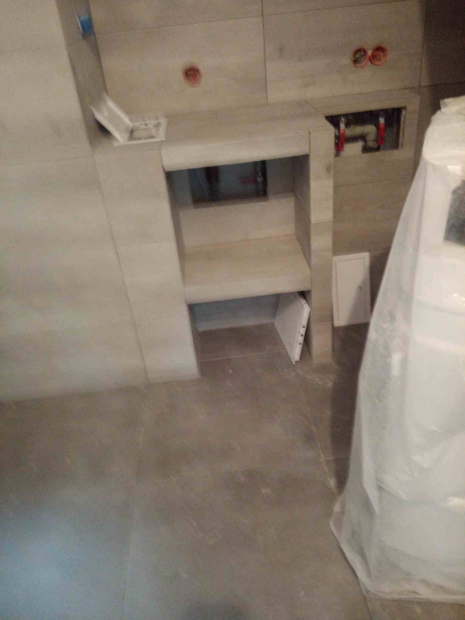 Niedokończona wnęka w ścianie wyłożonej płytkami, z widocznymi instalacjami hydraulicznymi i fragmentem otwartej klapy rewizyjnej, zabezpieczona folią malarską.