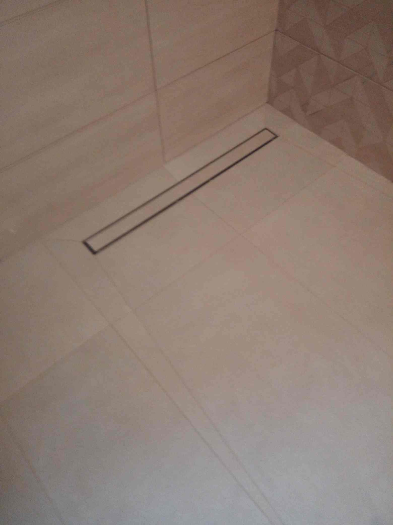 Beżowe płytki podłogowe i ścienne w kabinie prysznicowej z odpływem liniowym, widoczne fugi.