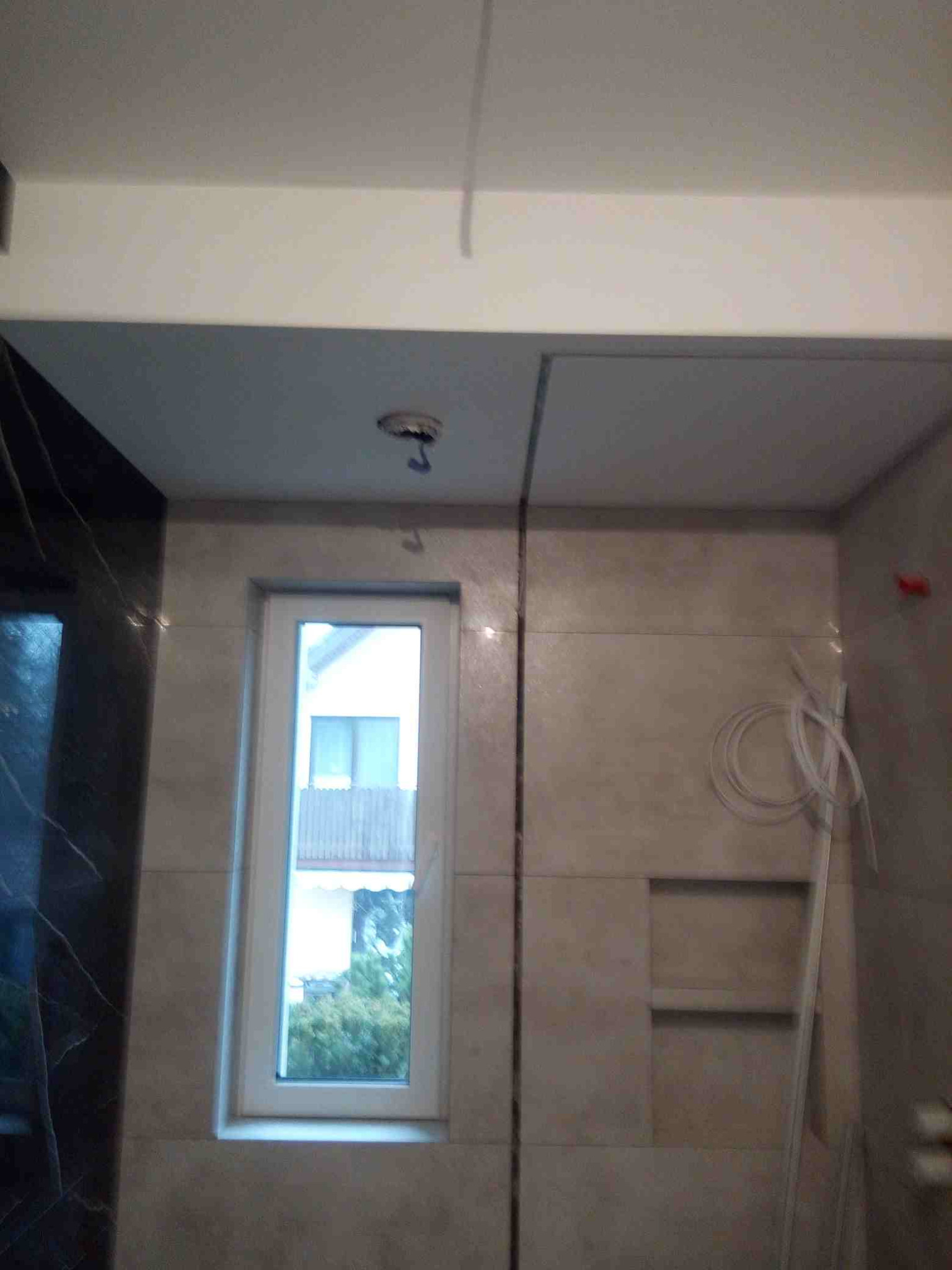 Wnętrze łazienki w trakcie wykańczania, widoczne beżowe płytki na ścianach, okno z widokiem na zieleń oraz przygotowane przewody elektryczne i otwarta półka w ścianie.