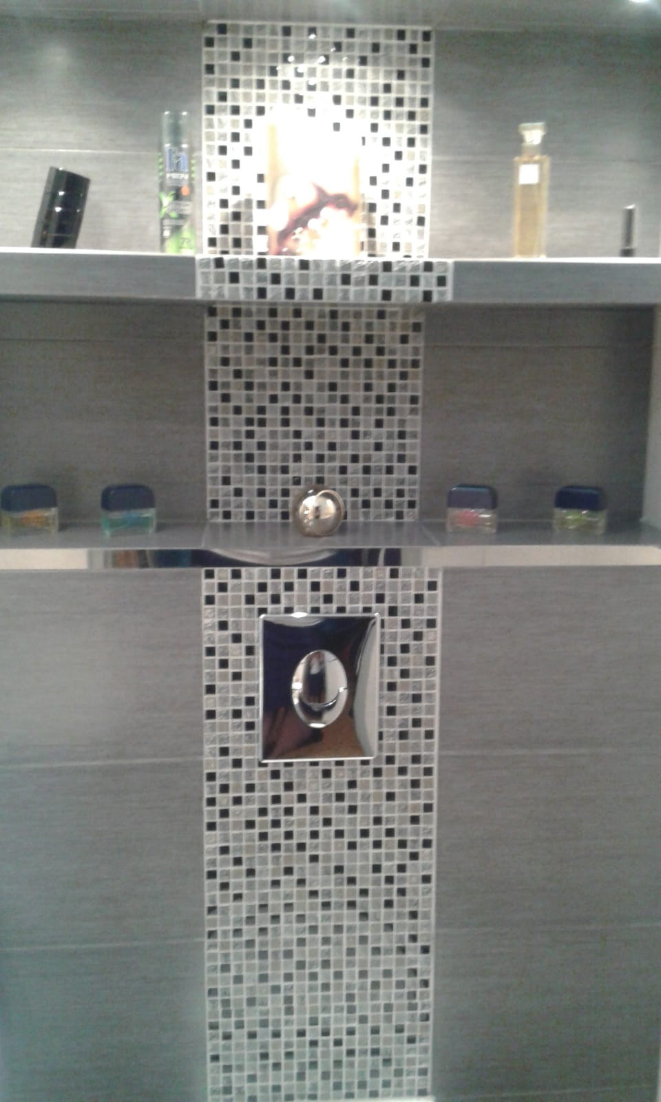 Wykończona ściana w łazience z szarymi płytkami i mozaiką z czarno-białych kwadratów, półki z kosmetykami i przycisk spłuczki WC w chromowanej ramce.