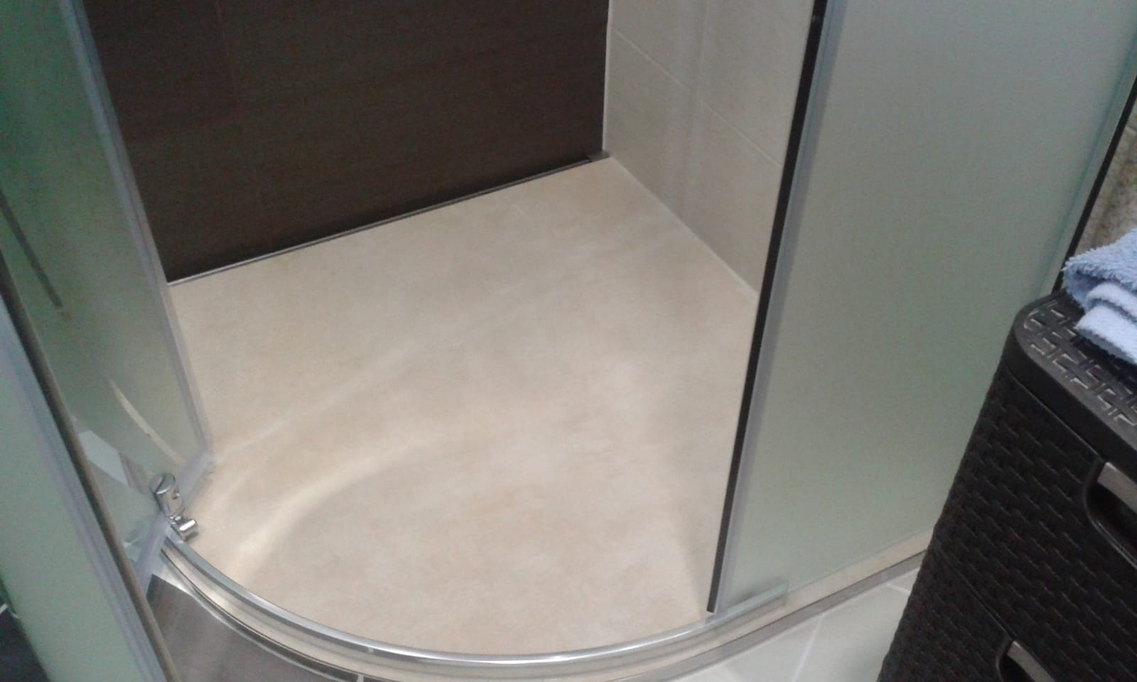 Kabina prysznicowa z beżowymi płytkami podłogowymi i brązową ścianą w tle, widoczny fragment kosza na pranie i ręcznik.