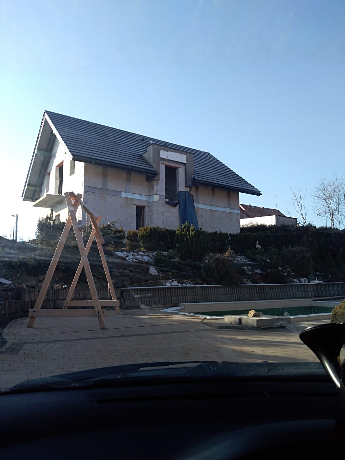 Niedokończony dom z szarej cegły z ciemnym dachem, widoczny zza drewnianej konstrukcji placu zabaw, teren wokół domu z elementami ogrodu.