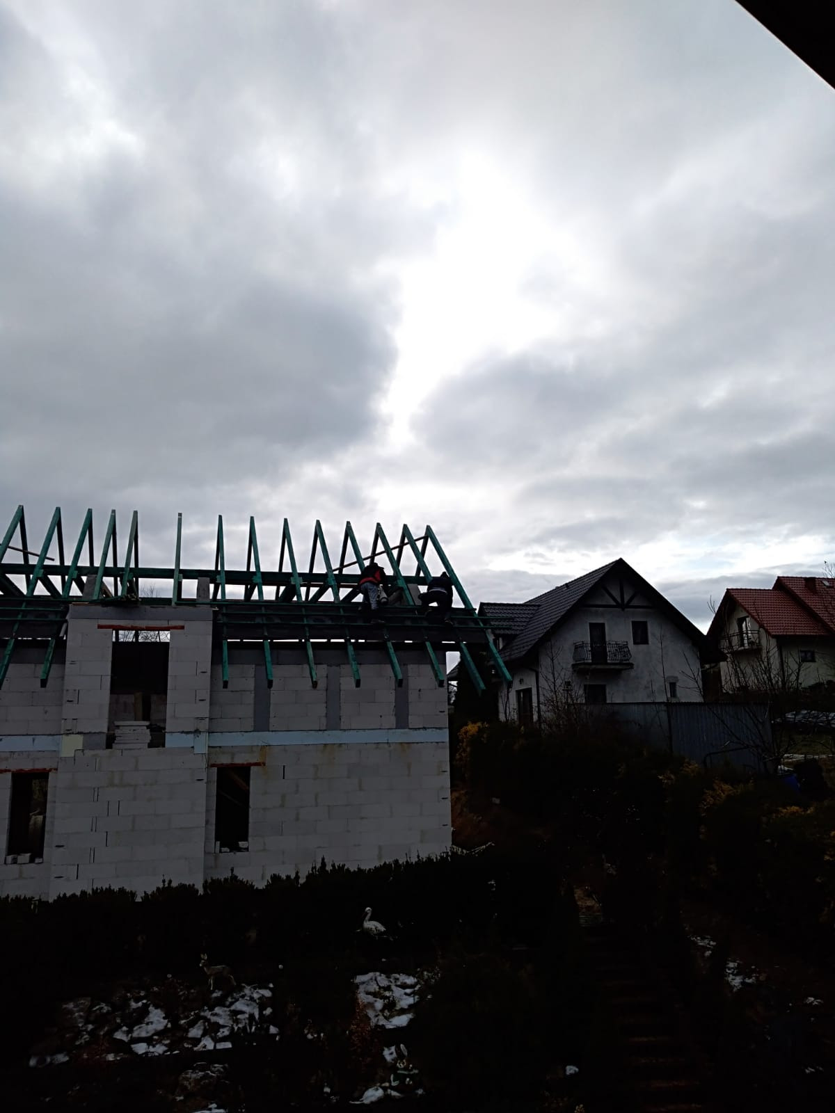 Budowa domu w stanie surowym otwartym, widoczne ściany z bloczków i drewniana konstrukcja dachu, dwóch robotników na dachu w pochmurny dzień.