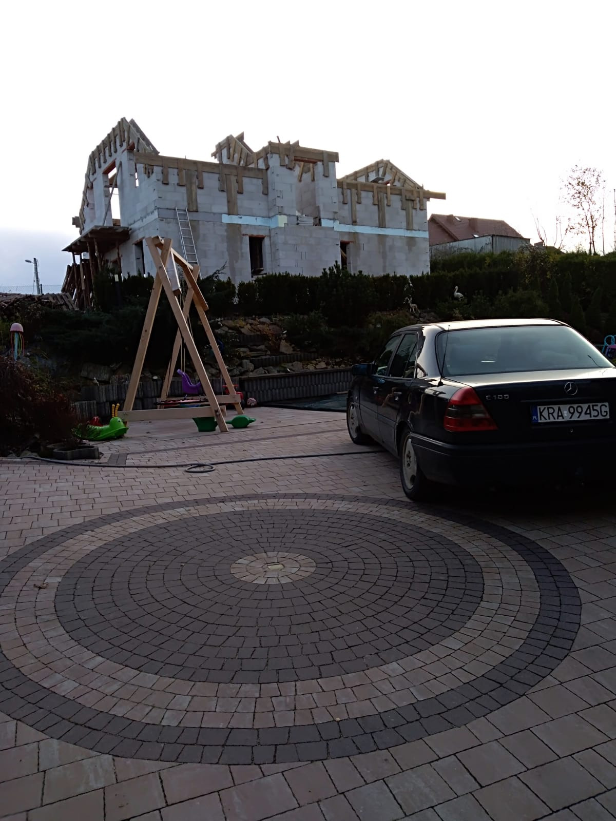Niedokończony budynek z białego betonu komórkowego z drewnianą konstrukcją dachu, widoczna drabina, plac zabaw dla dzieci i czarny samochód osobowy na posesji z kostki brukowej.