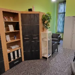 Biuro z nowoczesnymi meblami: drewniano-czarne szafy narożne z p&oacute;łkami, biała ażurowa szafka z kwiatem, szare fotele i metalowa szafa, widok zza biurka.