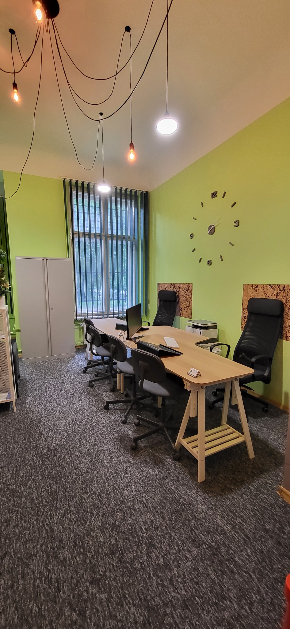 Biuro z zielonymi ścianami, dużym zegarem ściennym, stołem z krzesłami, lampami wiszącymi na kablach i szafą na dokumenty.