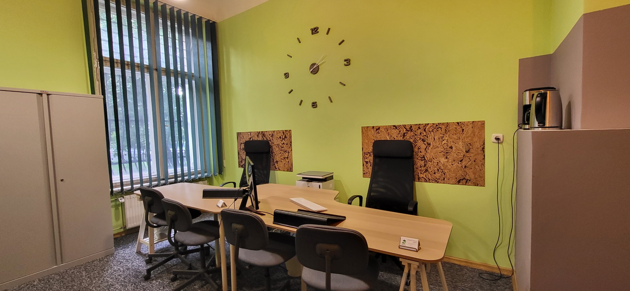 Jasne biuro z długim stołem konferencyjnym, krzesłami obrotowymi, dużym oknem z żaluzjami pionowymi, zegarem ściennym i ekspozycją korkową za krzesłami.