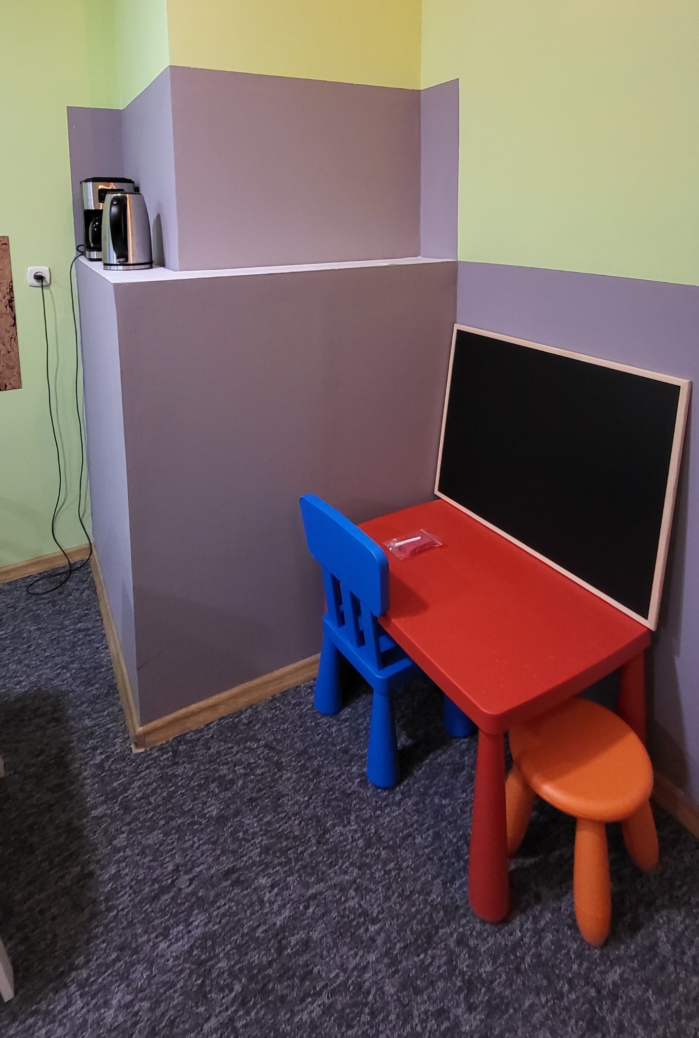 Pokój dziecięcy z małym czerwonym stolikiem, niebieskim krzesełkiem i pomarańczowym stołkiem, na stole leży paczka kredek, w tle czajnik i ekspres do kawy