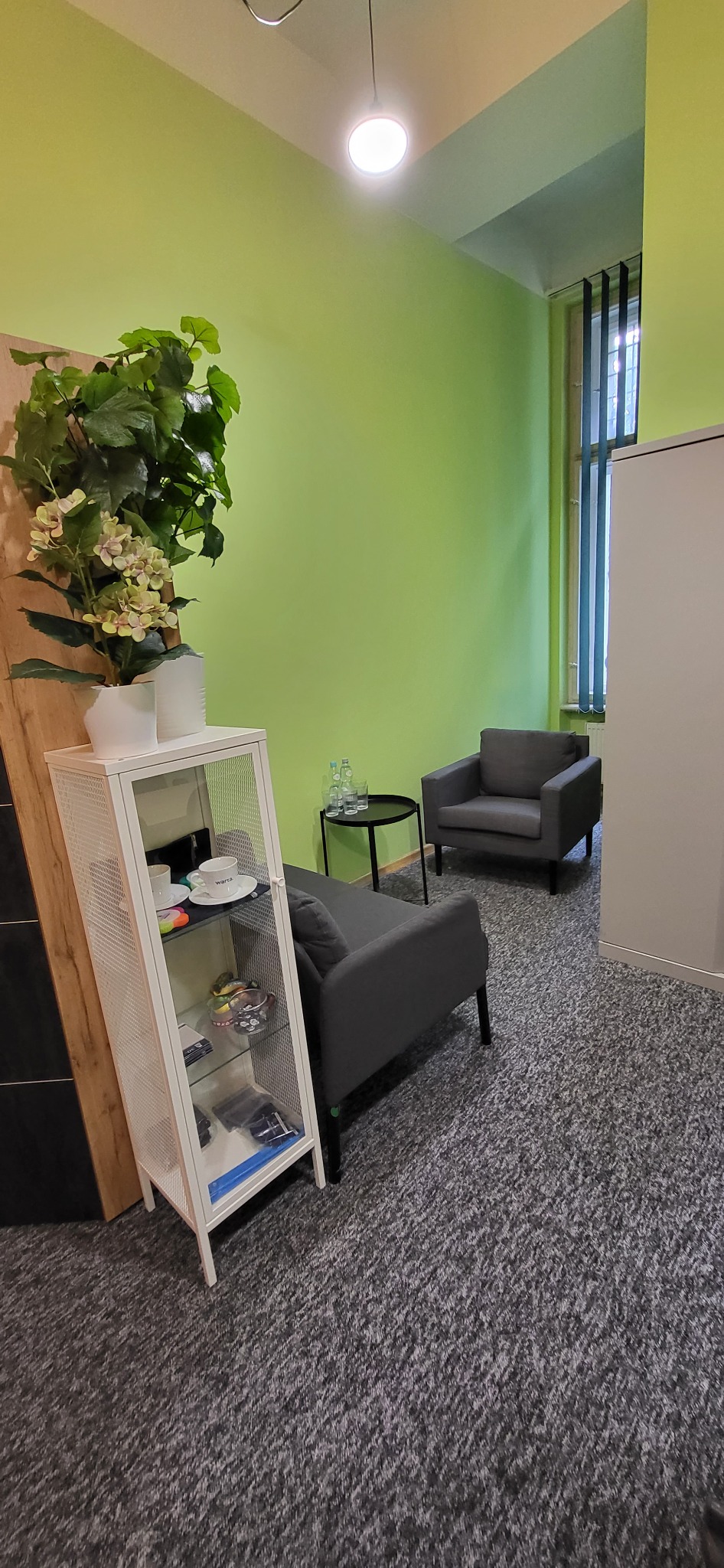 Jasnozielone biuro z dwoma szarymi fotelami, małym czarnym stolikiem z wodą i przeszkloną białą szafką z dekoracjami.