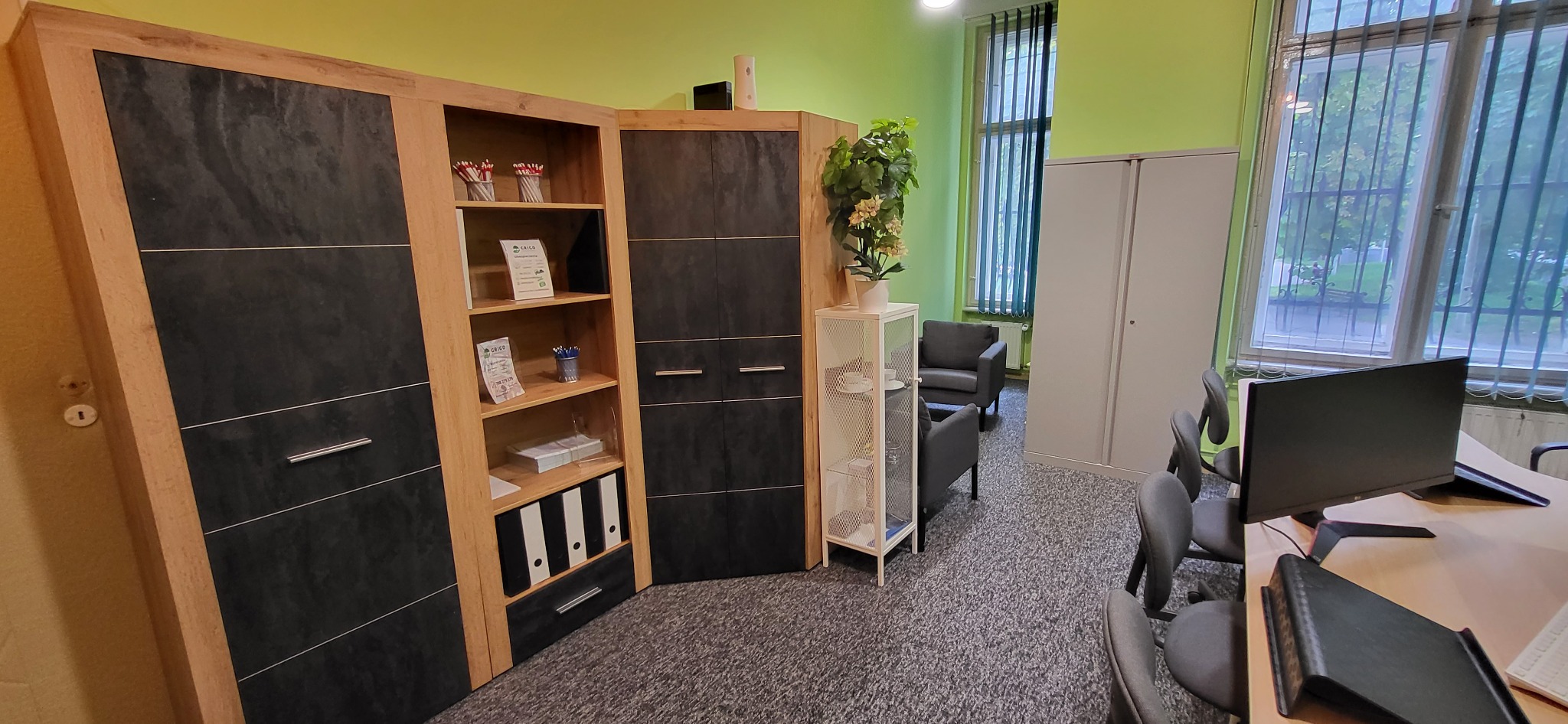 Biuro z nowoczesnymi meblami: drewniano-czarne szafy narożne z półkami, biała ażurowa szafka z kwiatem, szare fotele i metalowa szafa, widok zza biurka.