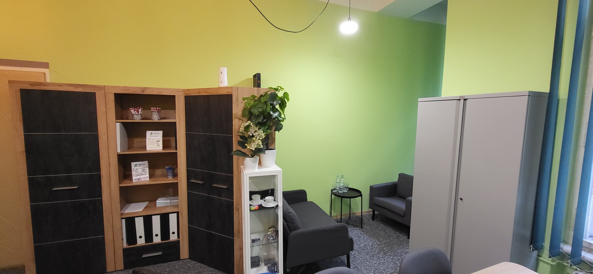 Jasnozielony gabinet z szarą sofą, fotelem, metalową szafą i regałem z dokumentami, dwie butelki wody na stoliku kawowym.