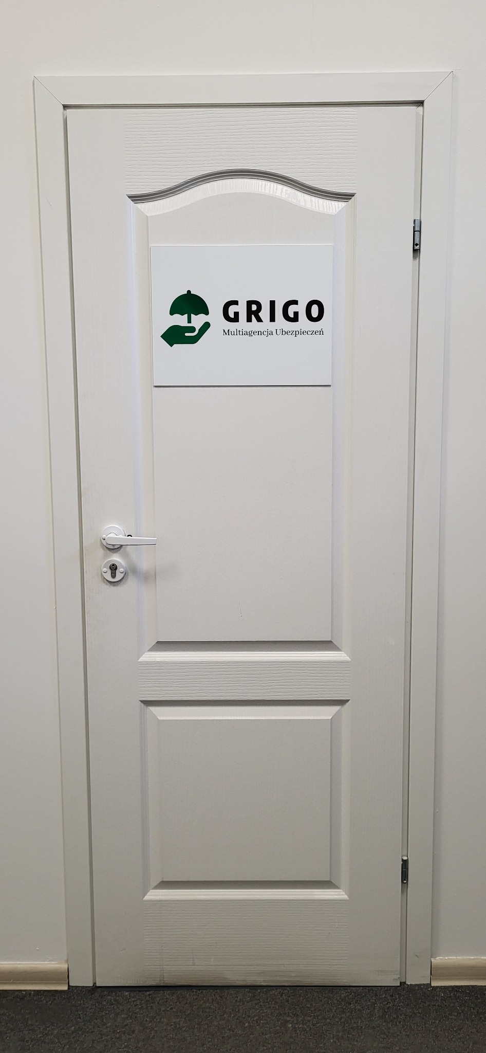 Białe drzwi z tabliczką informacyjną multiagencji ubezpieczeniowej Grigo, z logo przedstawiającym dłoń trzymającą parasol, znajdujące się w biurze.