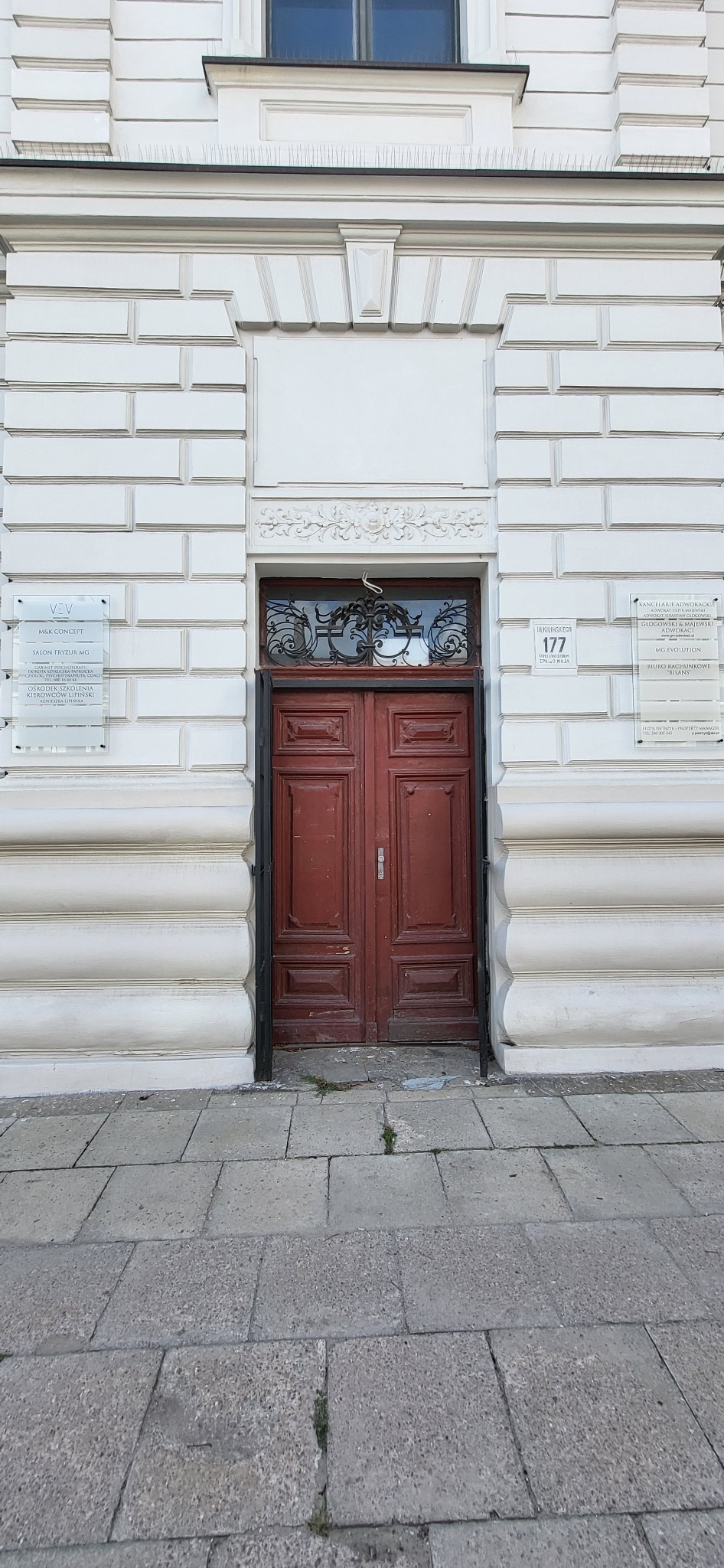 Wejście do budynku z drewnianymi drzwiami w kolorze bordo, nad którymi widnieje ozdobna krata, a obok tabliczki informacyjne z nazwami firm i numerem adresu 177.