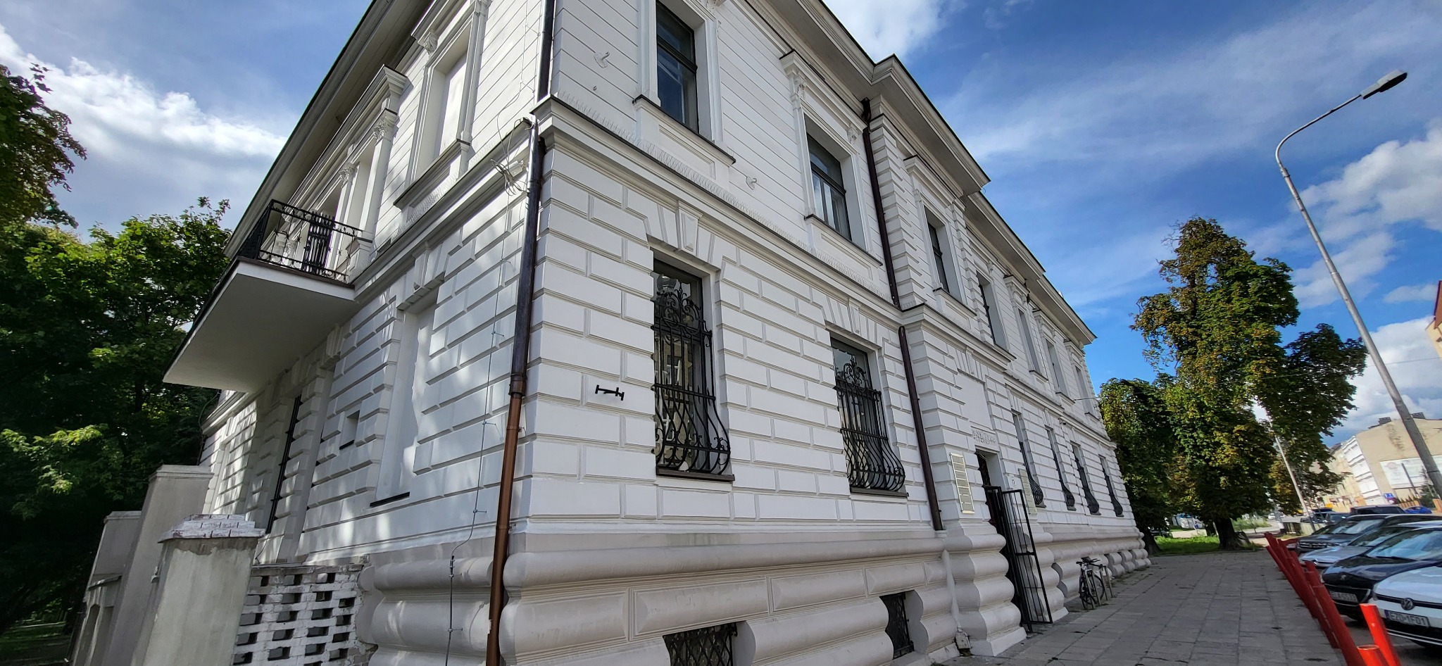 Elewacja odnowionego, dwupiętrowego budynku z balkonem i ozdobnymi kratami w oknach, widoczna z poziomu chodnika, na tle błękitnego nieba z chmurami.