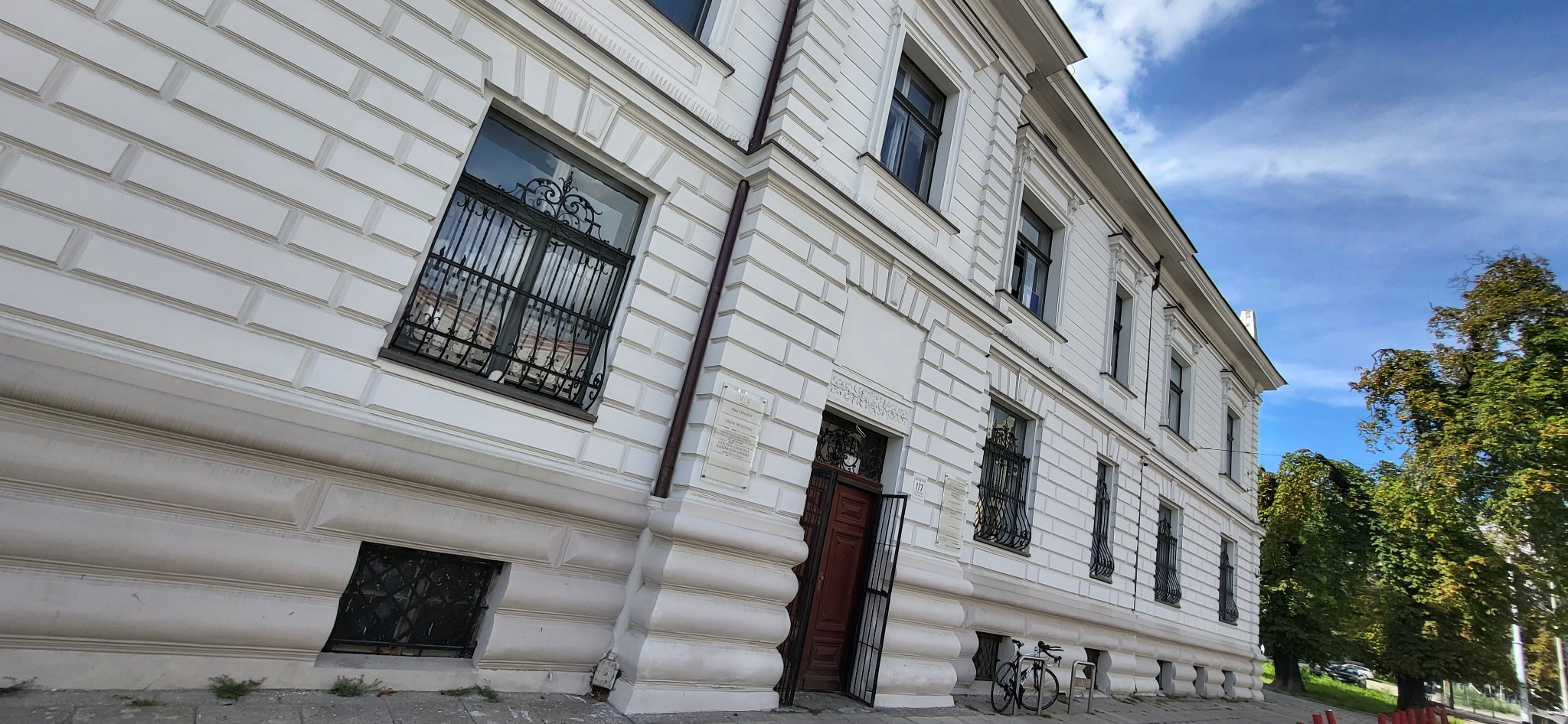 Elewacja odnowionego, białego budynku z dekoracyjnymi oknami i żeliwnymi kratami, rower oparty o stojak przed wejściem.