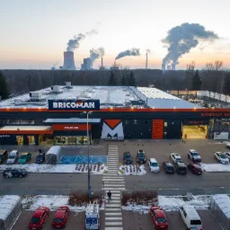Panoramiczne ujęcie sklepu budowlanego Bricoman z parkingiem, widok z góry, w tle kominy elektrowni i zachodzące słońce.